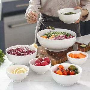 Person preparing a salad with various ingredients in white bowls on a kitchen counter.