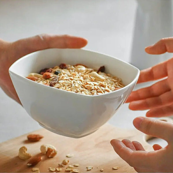 White bowl filled with cereal and nuts on a wooden surface, with hands reaching towards it.