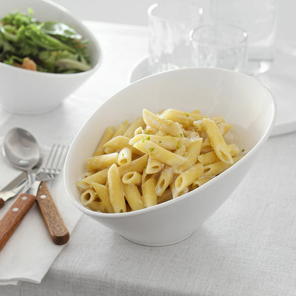 White bowl of pasta on a table with cutlery and another dish in the background