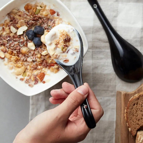 Hand holding a Black spoon with yogurt and granola over a bowl of the same, on a checkered tablecloth.