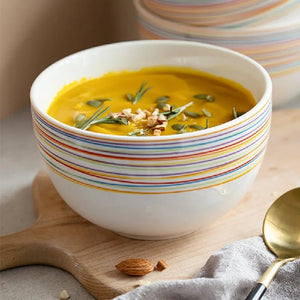 White bowl with colorful stripes filled with yellow soup, garnished with herbs and nuts, on a wooden board.