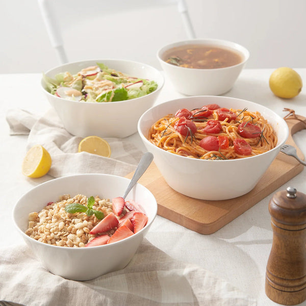 Four white bowls with different dishes on a light surface with a lemon and wooden pepper grinder.