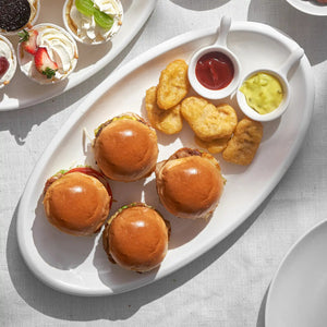 ceramic serving platter of mini burgers and tater tots with side dishes on a light gray surface