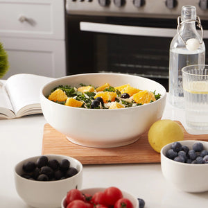 Salad in a white bowl on a kitchen counter with fruits and a water bottle in the background.