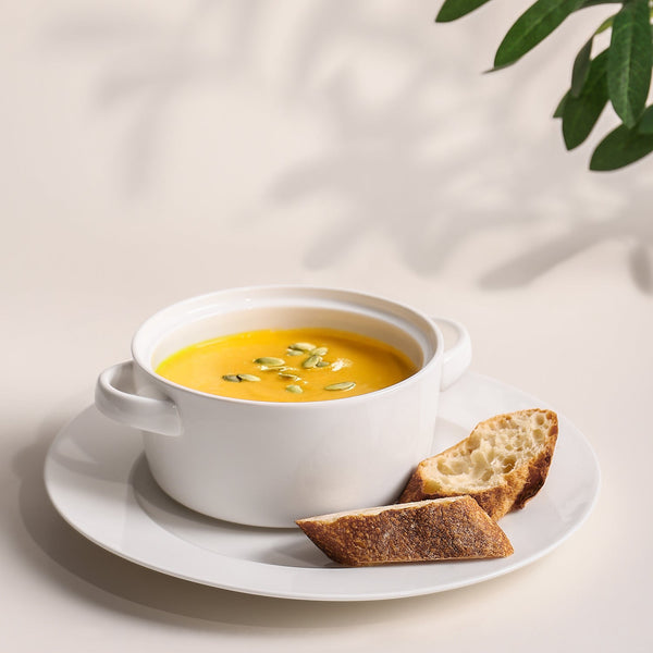 White bowl of yellow soup with bread on a white plate, light background