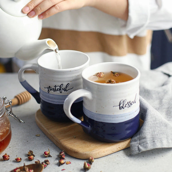Two mugs with 'grateful' and 'blessed' text being filled with a liquid on a wooden board.