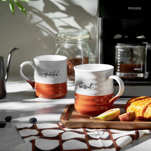 Two 18 oz mugs with 'grateful' and 'blessed' text on a kitchen counter with a coffee maker and fruit.