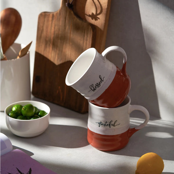 Two orange coffee mugs with 'blessed' and 'grateful' text on a kitchen counter.