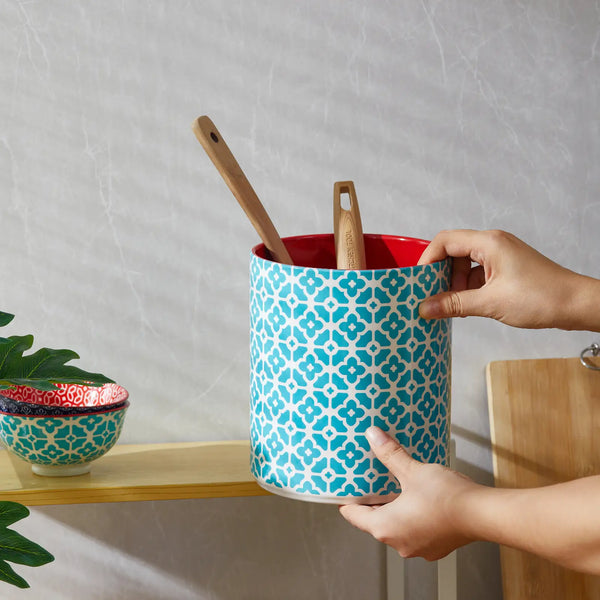 Person holding a blue patterned utensil holder with red interior, wooden spoons inside, on a neutral background.