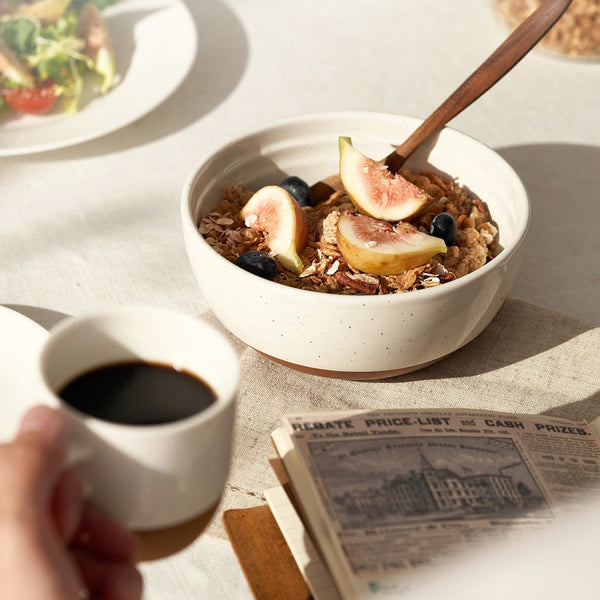 Bowl of cereal with figs and blueberries, cup of coffee, and newspaper on a table.