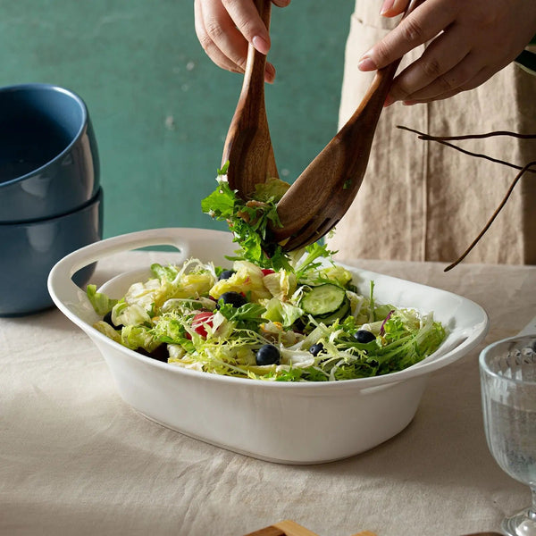 Person tossing a salad in a white bowl with wooden utensils on a table.