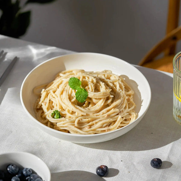 White bowl of pasta with a glass of water on a light surface