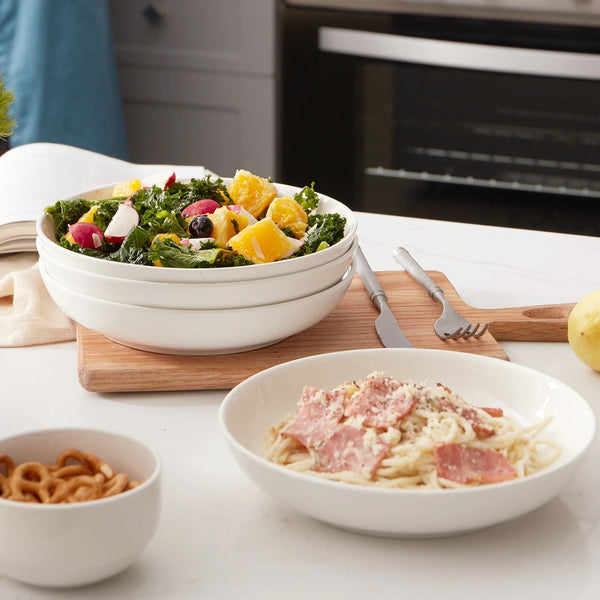 Bowl of salad with fruit and vegetables on a kitchen counter with pasta dish and snacks.