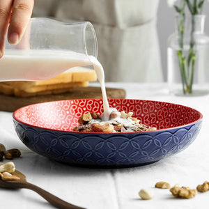 Person pouring milk into a blue and red bowl with cereal and nuts on a light background