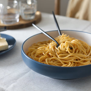 Bowl of spaghetti on a table with a plate of cheese and glasses in the background
