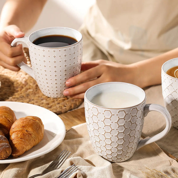 Person holding a patterned mug with coffee, surrounded by croissants and another mug on a textured surface.