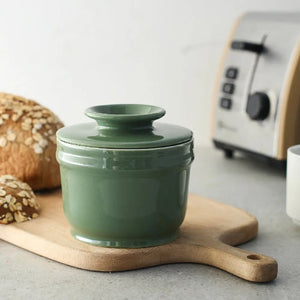 Green ceramic container with a lid on a wooden cutting board next to bread and a toaster.