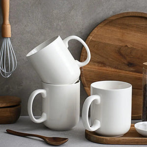 white porcelain coffee mugs stacked on a gray surface with wooden utensils and a wooden cutting board in the background.