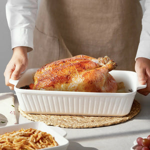 Person holding a white dish with a roasted chicken on a table with side dishes.