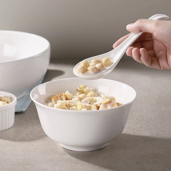 White bowl with cereal and a hand holding a white spoon with cereal, on a neutral background.