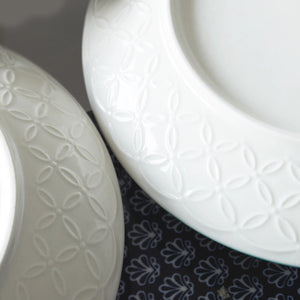 Two white ceramic plates with embossed patterns on a dark fabric background