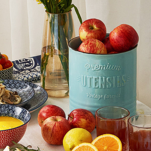 Teal container with apples on a table with fruit and glasses.