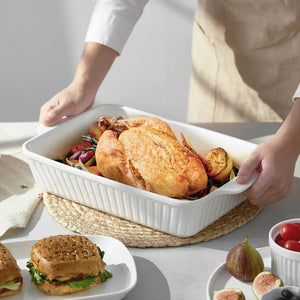 Person holding a white ceramic roasting dish with roasted chicken and vegetables on a table.