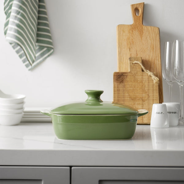 Green ceramic baking dish on a kitchen counter with cutting board and utensils in the background